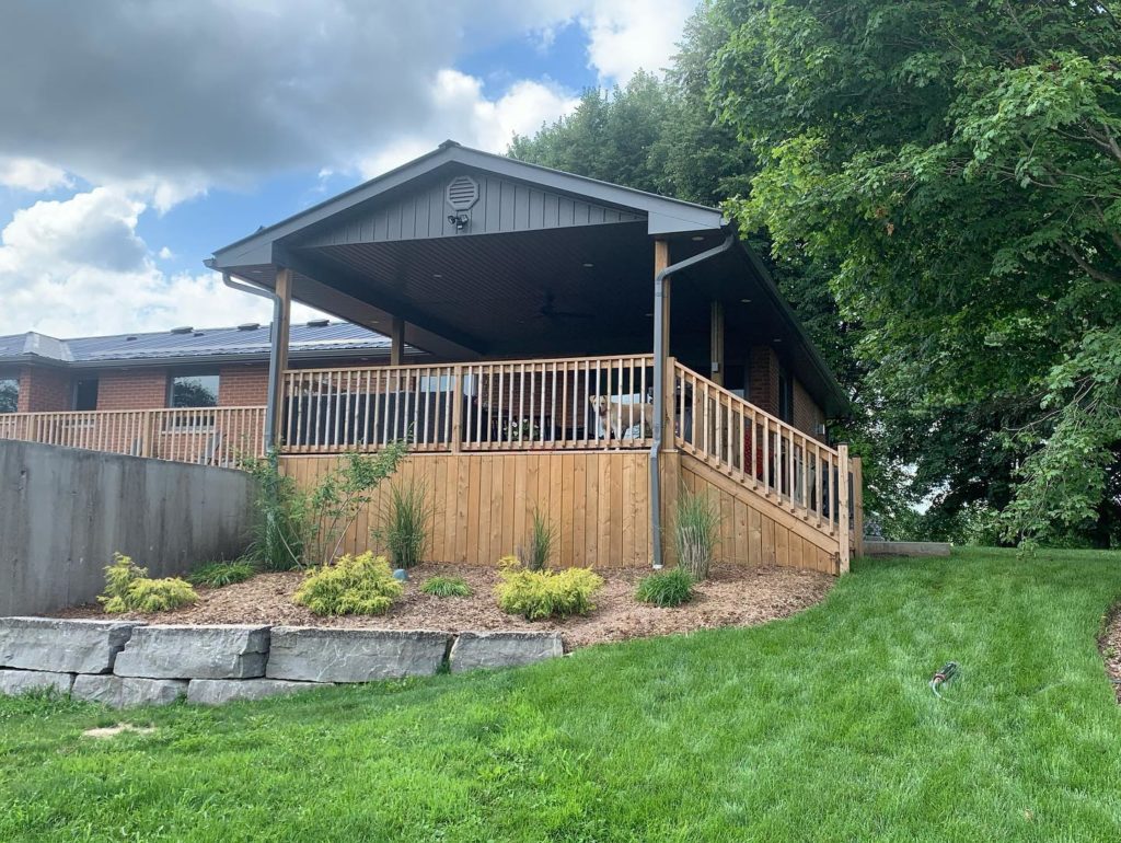 Wooden deck with small garden in front surrounded by large patch of grass with dog on the deck